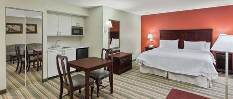 A hotel room with a bed, a desk with a chair, a small table, a mirror, a microwave, and a sink.
