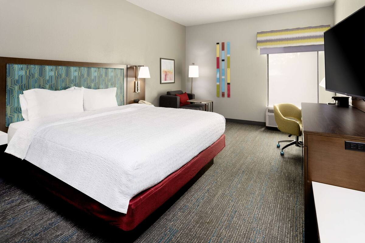A hotel room with a large bed, a desk with a computer, a chair, a desk lamp, a telephone, and a TV.