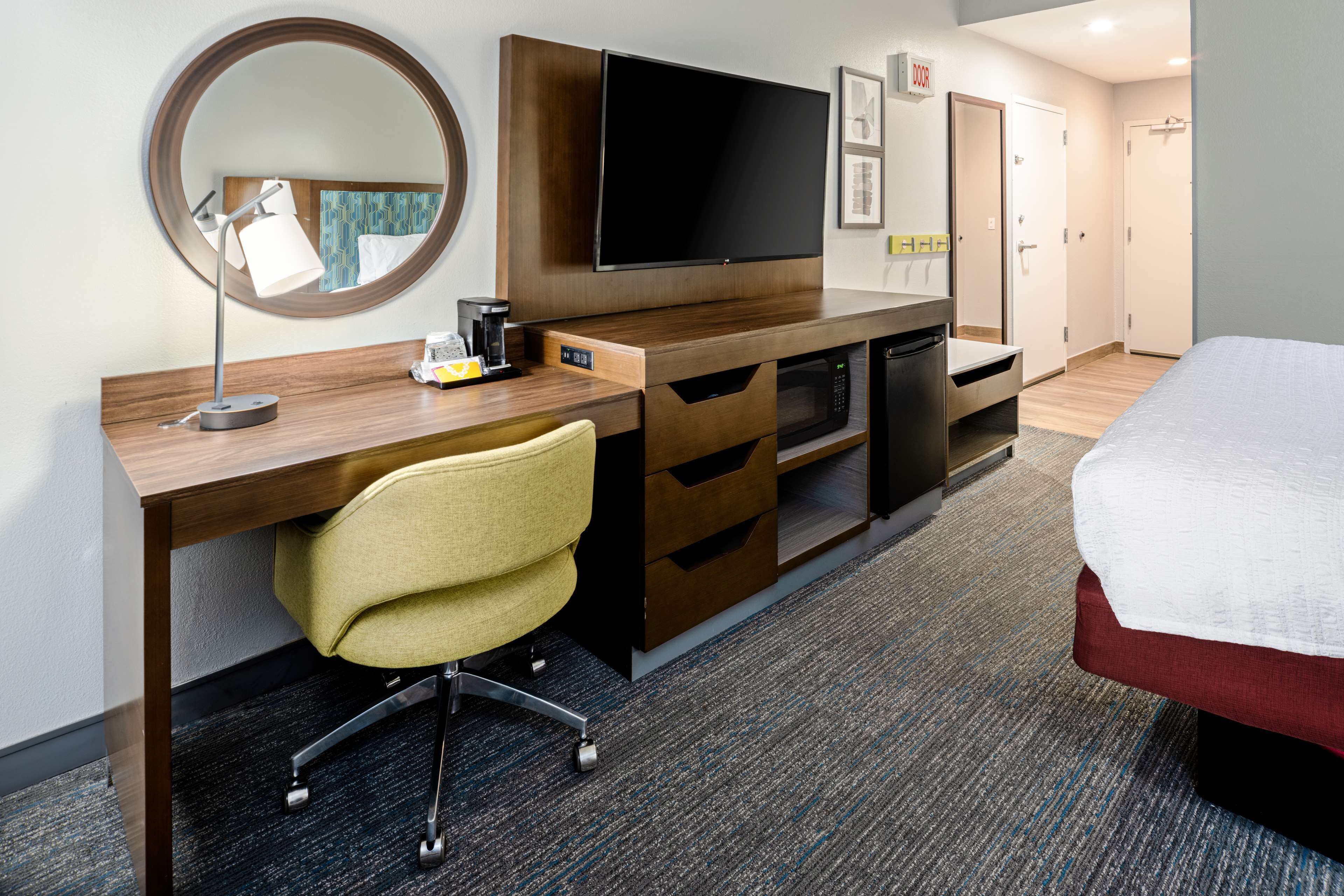 A hotel room with a wooden desk, a television, a mirror, a chair, a bed, and a wardrobe.