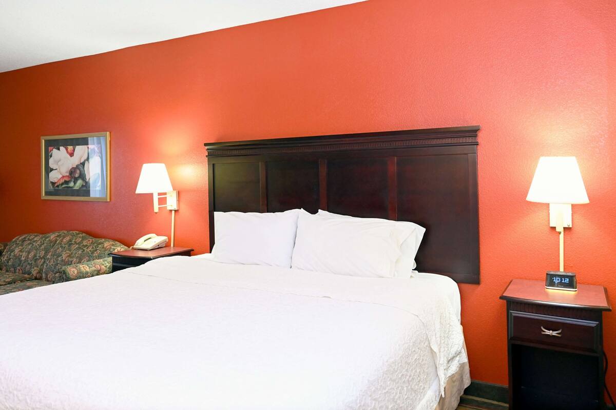 A hotel room with a large bed, two bedside tables with lamps, a nightstand with a clock, and a floral-patterned couch.