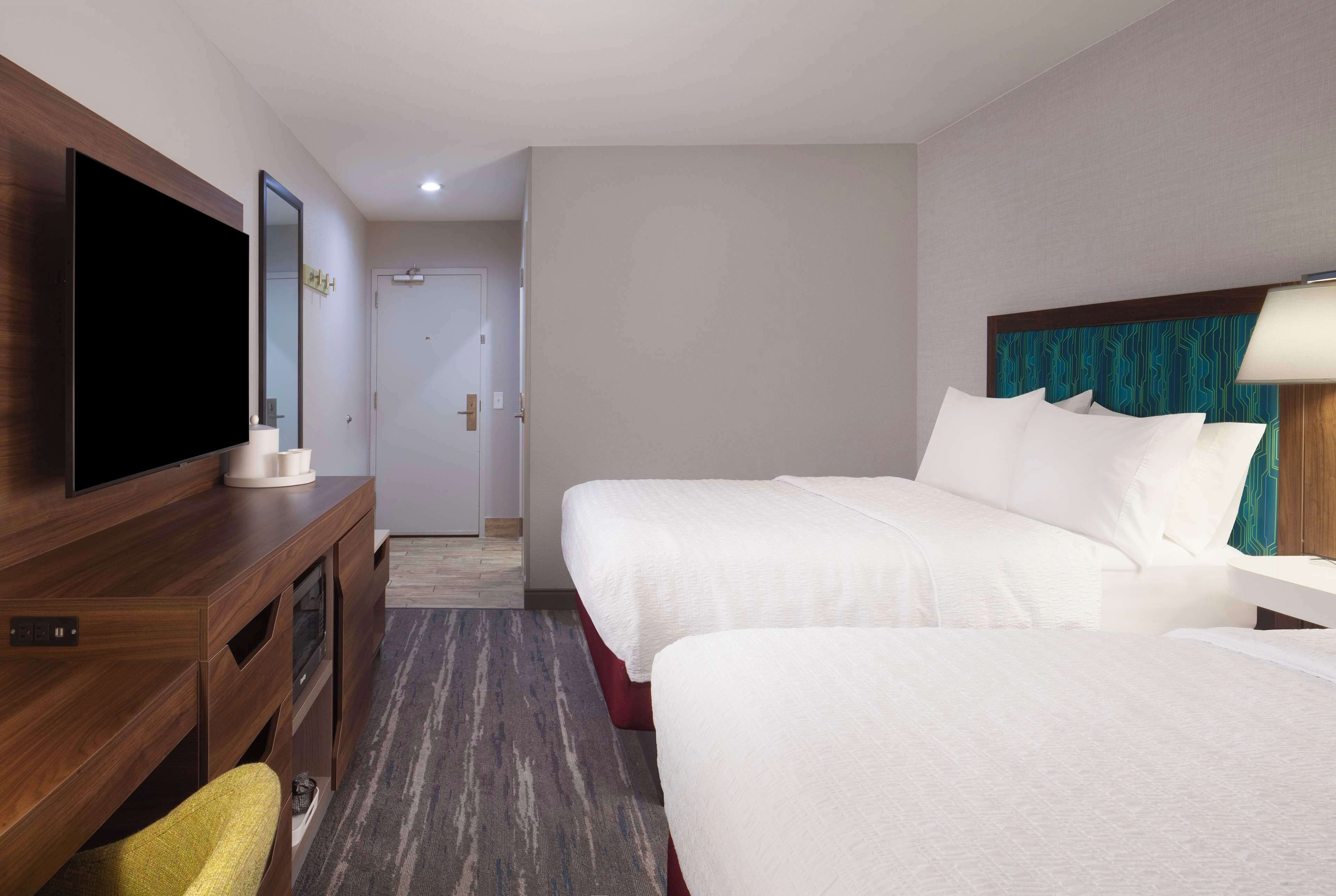 Quarto de hotel com televisão sobre um móvel de madeira, duas camas com roupa de cama branca, cabeceira de madeira e chão coberto por carpete.