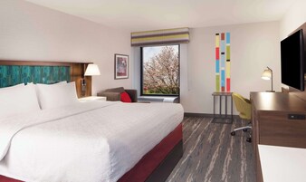 A hotel room with a large bed, a desk with a TV, a chair, a window with a view of trees, and a colorful wall decoration.