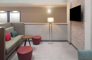 Una habitación de hotel moderna con un sofá, dos otomanos rojos, una lámpara de pie y un televisor de pantalla plana montado en la pared.