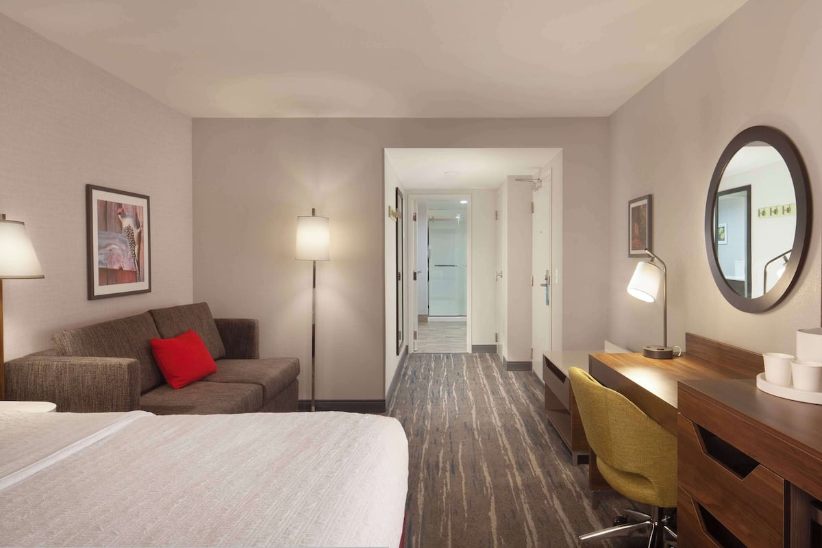 Habitación de hotel con cama, escritorio, silla, lámpara, espejo y un cuadro en la pared.
