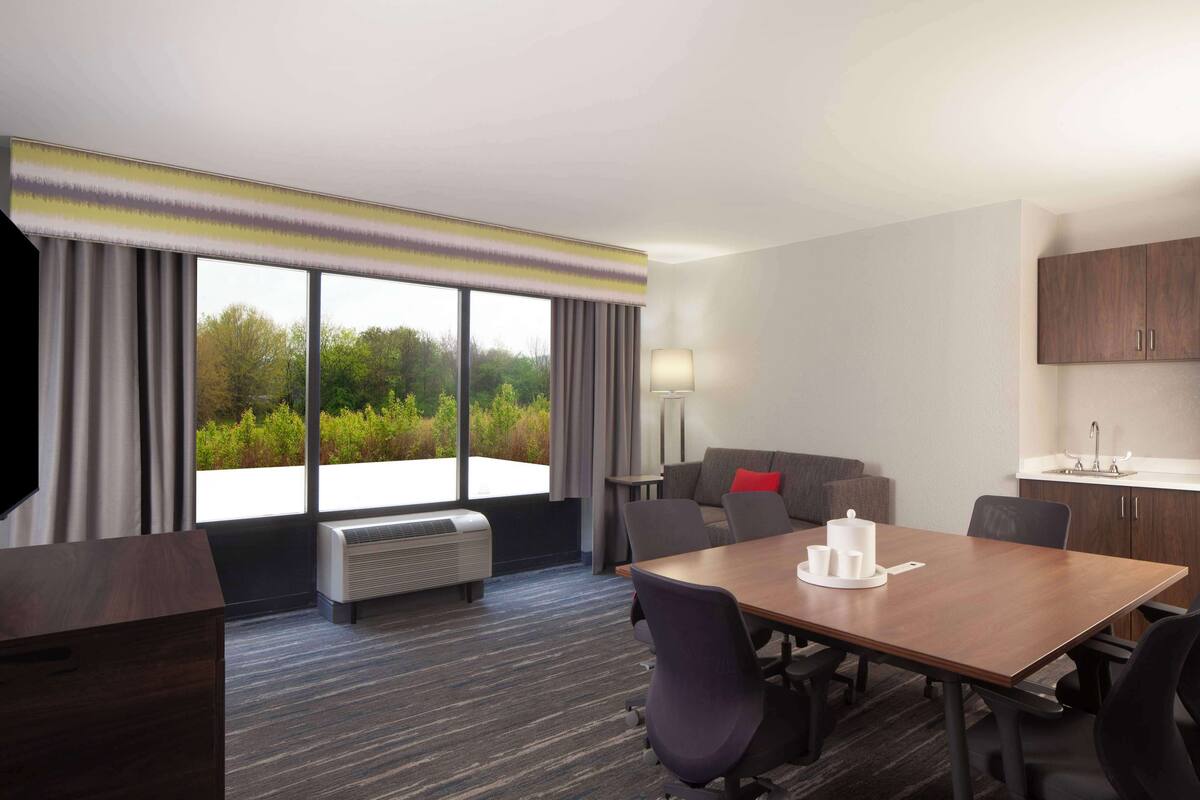 Habitación de hotel con un ventanal grande, un sofá, una mesa de comedor con sillas, una kitchenette y un televisor de pantalla plana.