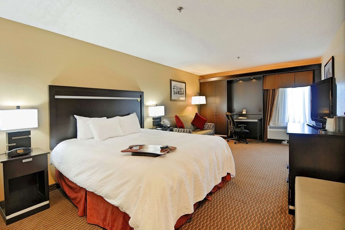 A hotel room with a large bed, a desk, a chair, a TV, and a desk lamp.