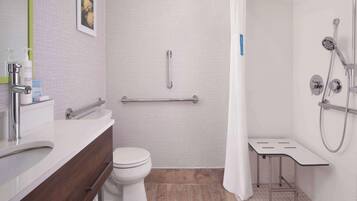 Une salle de bain moderne avec des toilettes blanches, un lavabo avec un meuble-vasque en bois, une douche équipée d’un pommeau de douche à main et d’une barre d’appui.