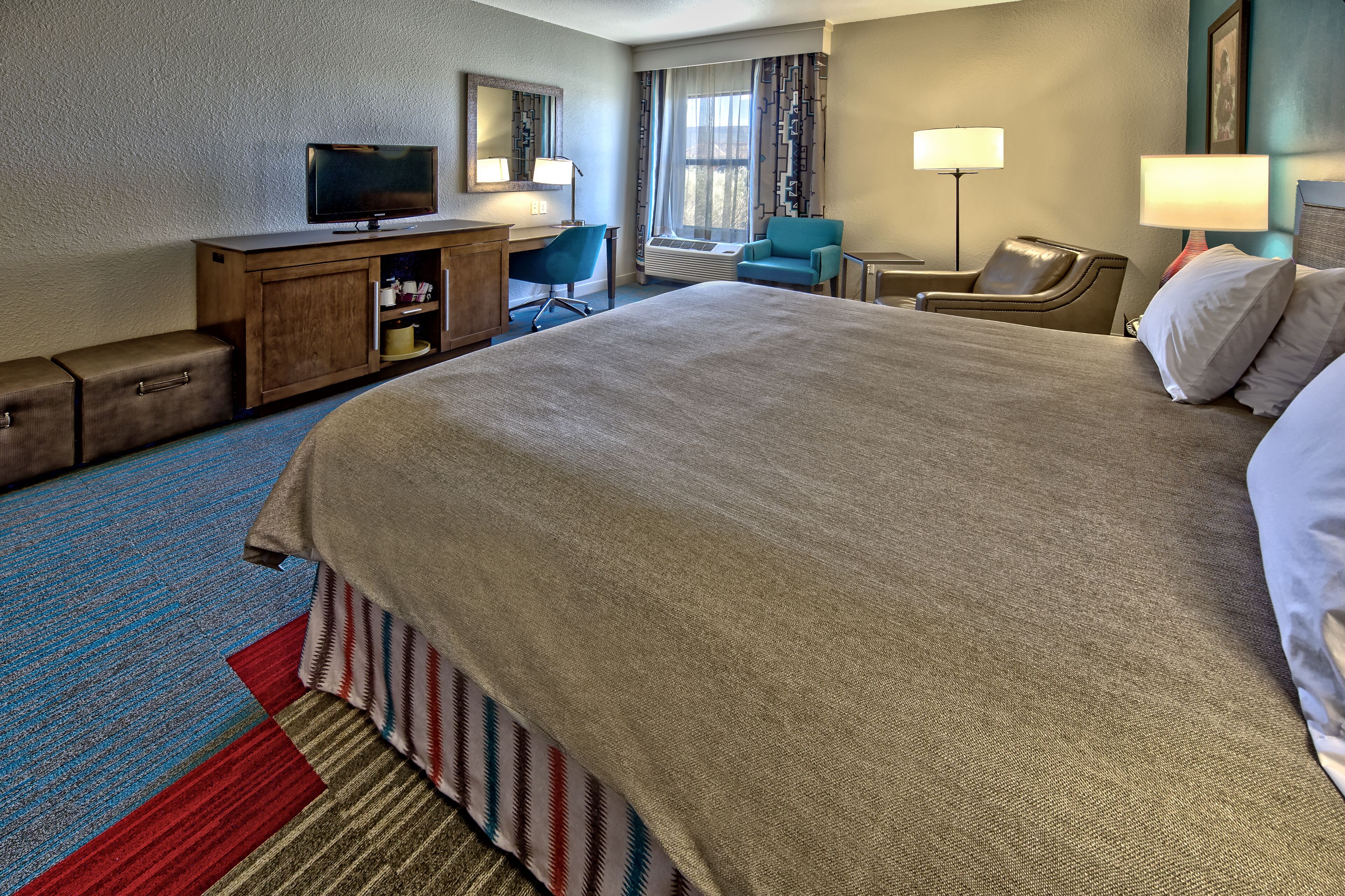 A hotel room with a large bed, a desk with a television, a chair, a desk lamp, and a window with curtains.