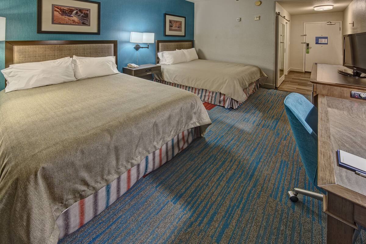 A hotel room with two beds, a desk with a chair, a TV, and a bathroom visible in the background.