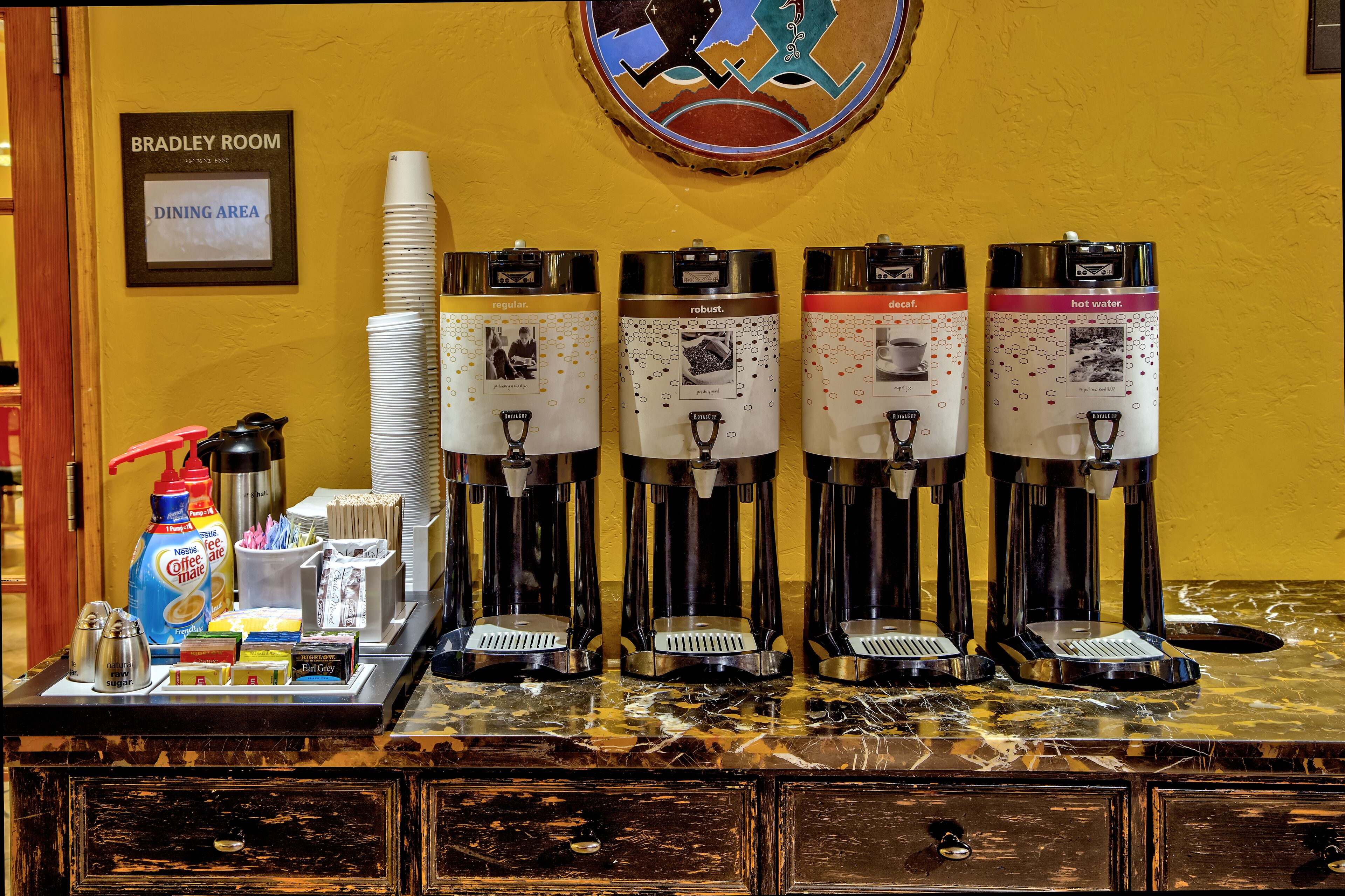 A coffee station with four dispensers labeled 'regular,' 'robust,' 'decaf,' and 'hot water.' There is a sign above indicating 'Bradley Room' and 'Dining Area.'