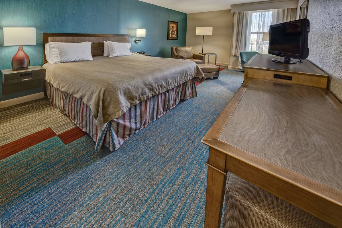 A hotel room with a large bed, a desk with a television, a chair, and a nightstand with a lamp.