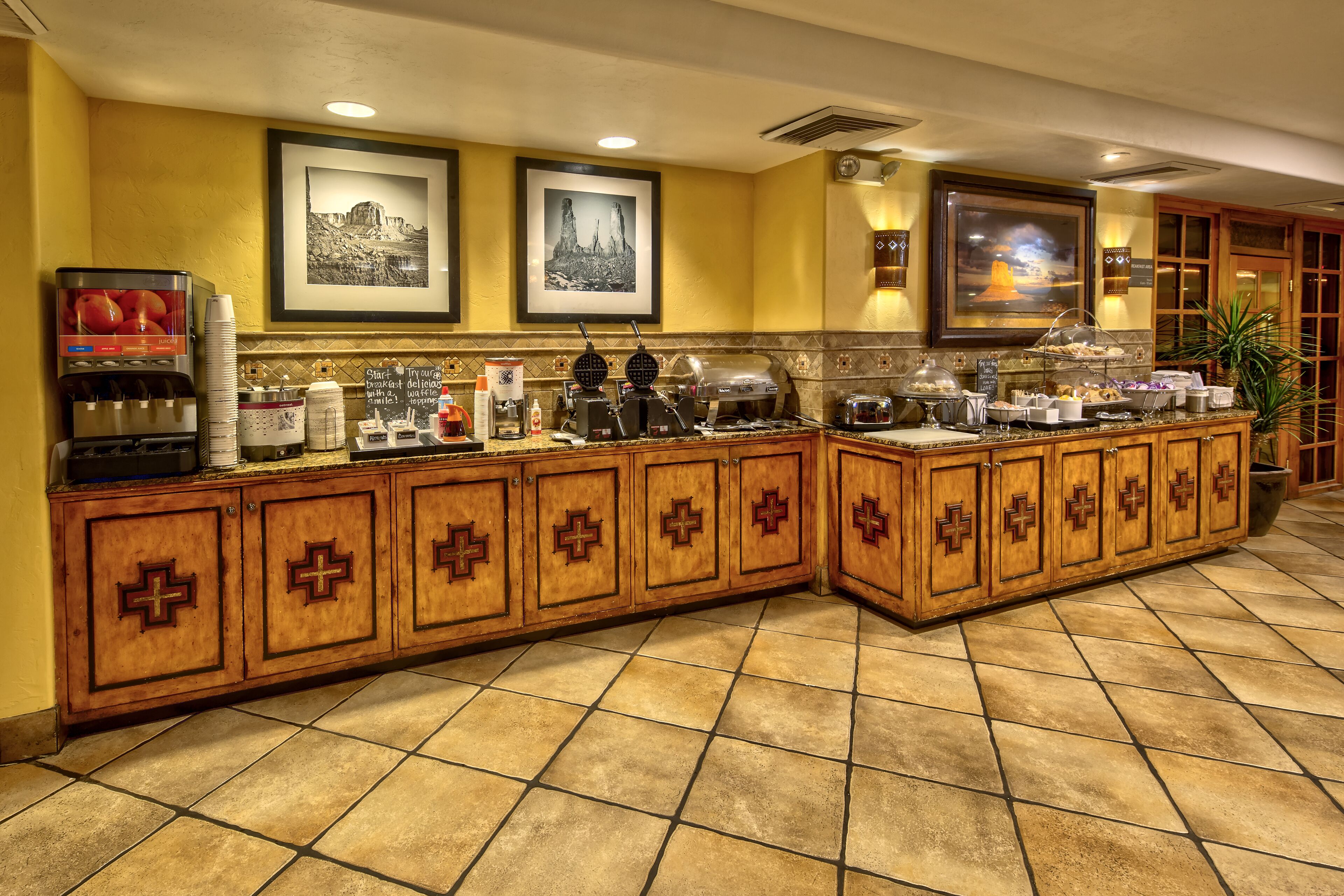 A hotel breakfast bar with a coffee machine, toaster, and various food items. There are framed pictures on the wall and a potted plant in the corner.
