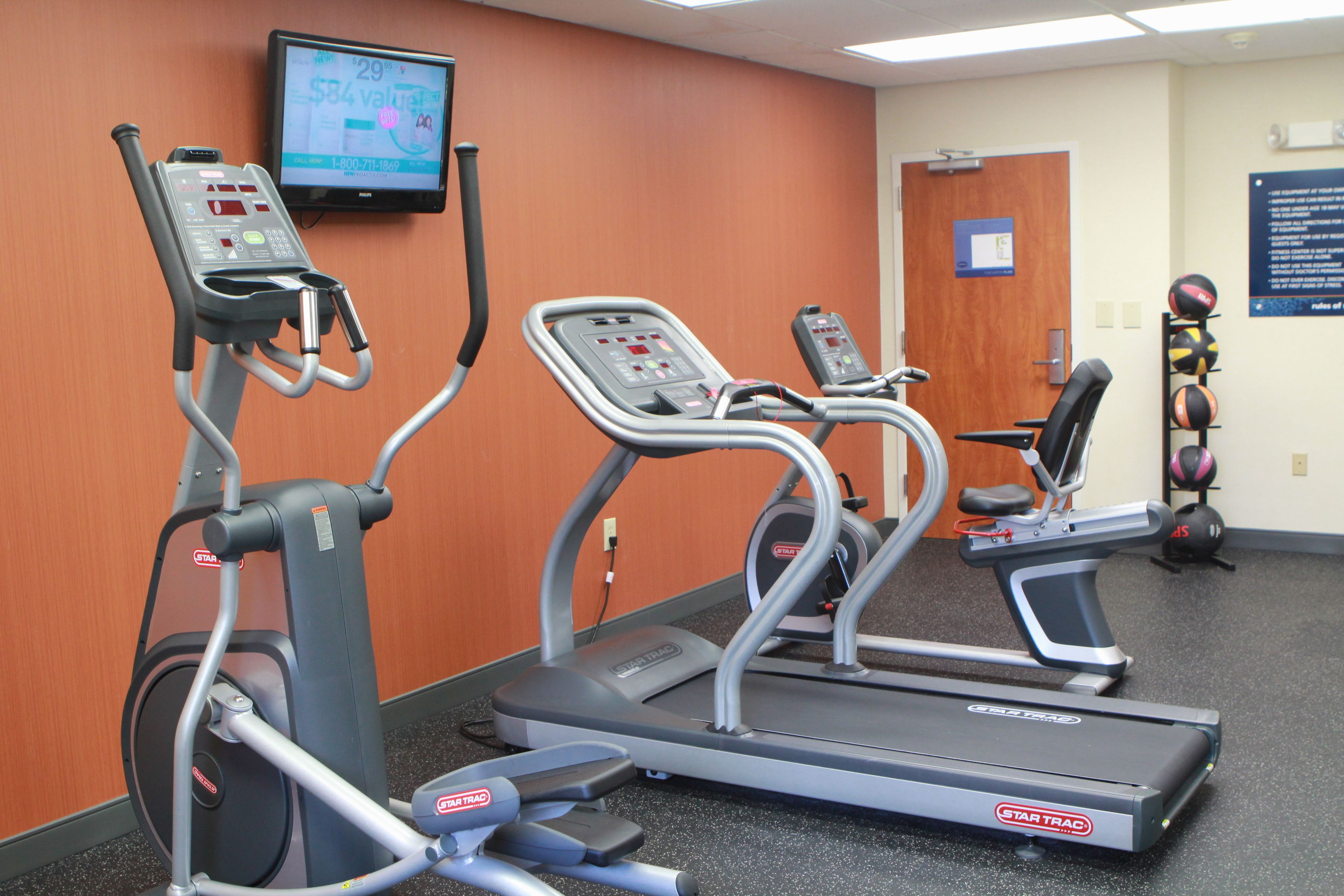 A fitness center with treadmills and elliptical machines, a television displaying workout information, and a wall-mounted information board.