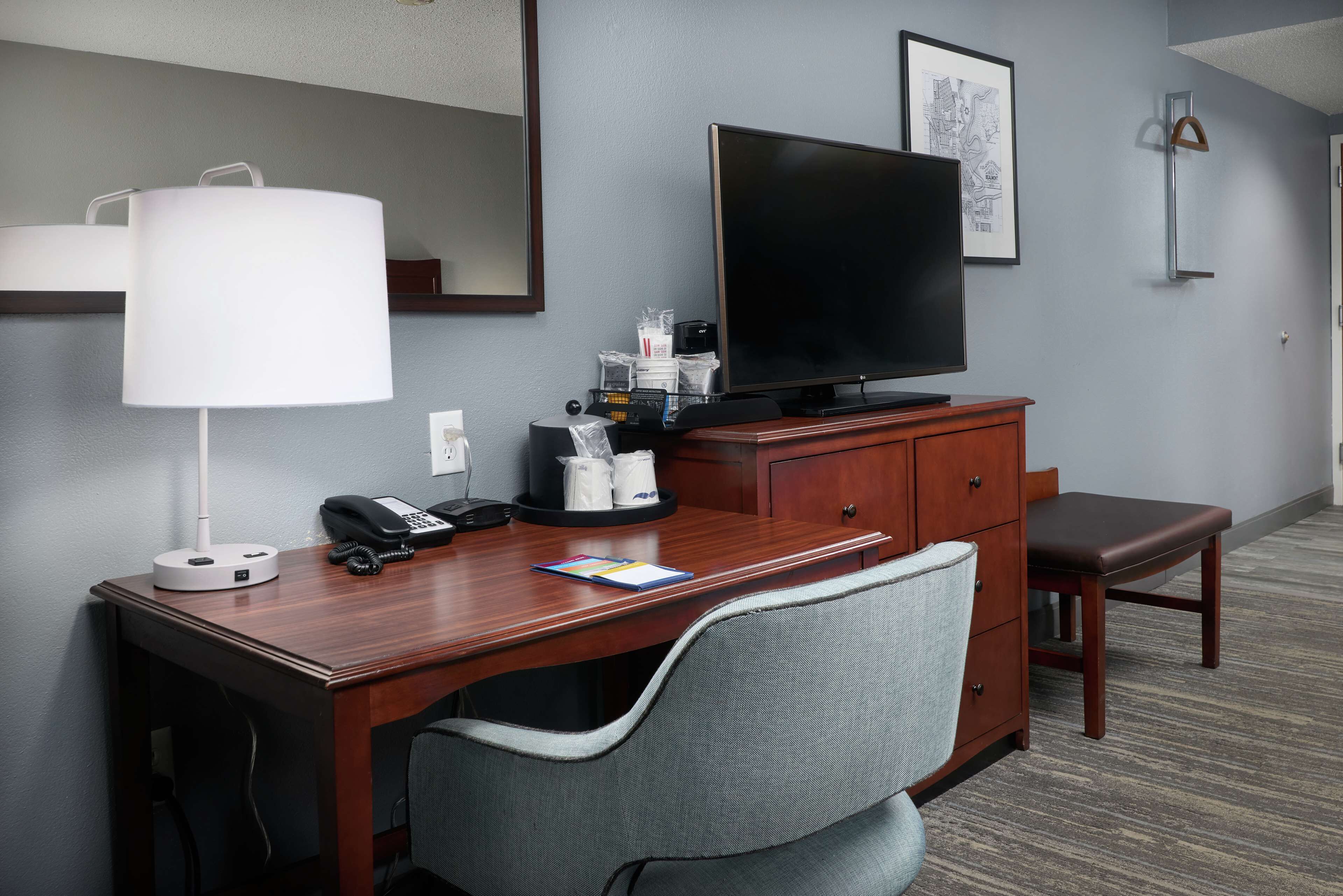 A hotel room with a wooden desk, a television, a lamp, a telephone, a coffee maker, and a chair.