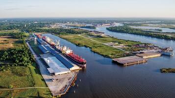 An industrial port with cargo ships docked, a large warehouse, and a river with surrounding greenery.
