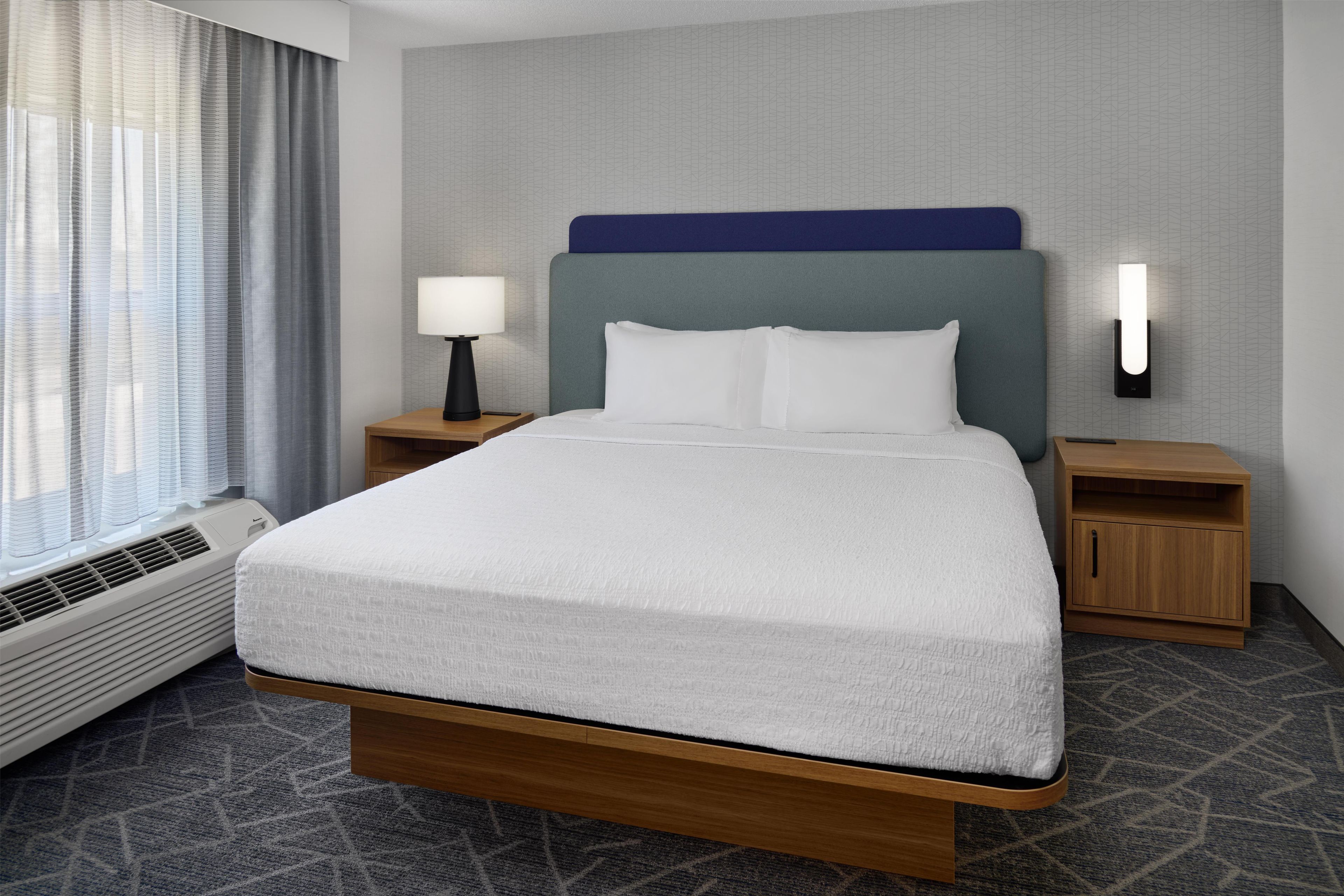 A hotel room with a bed, bedside tables, a lamp, a wall-mounted light, and a window with curtains.