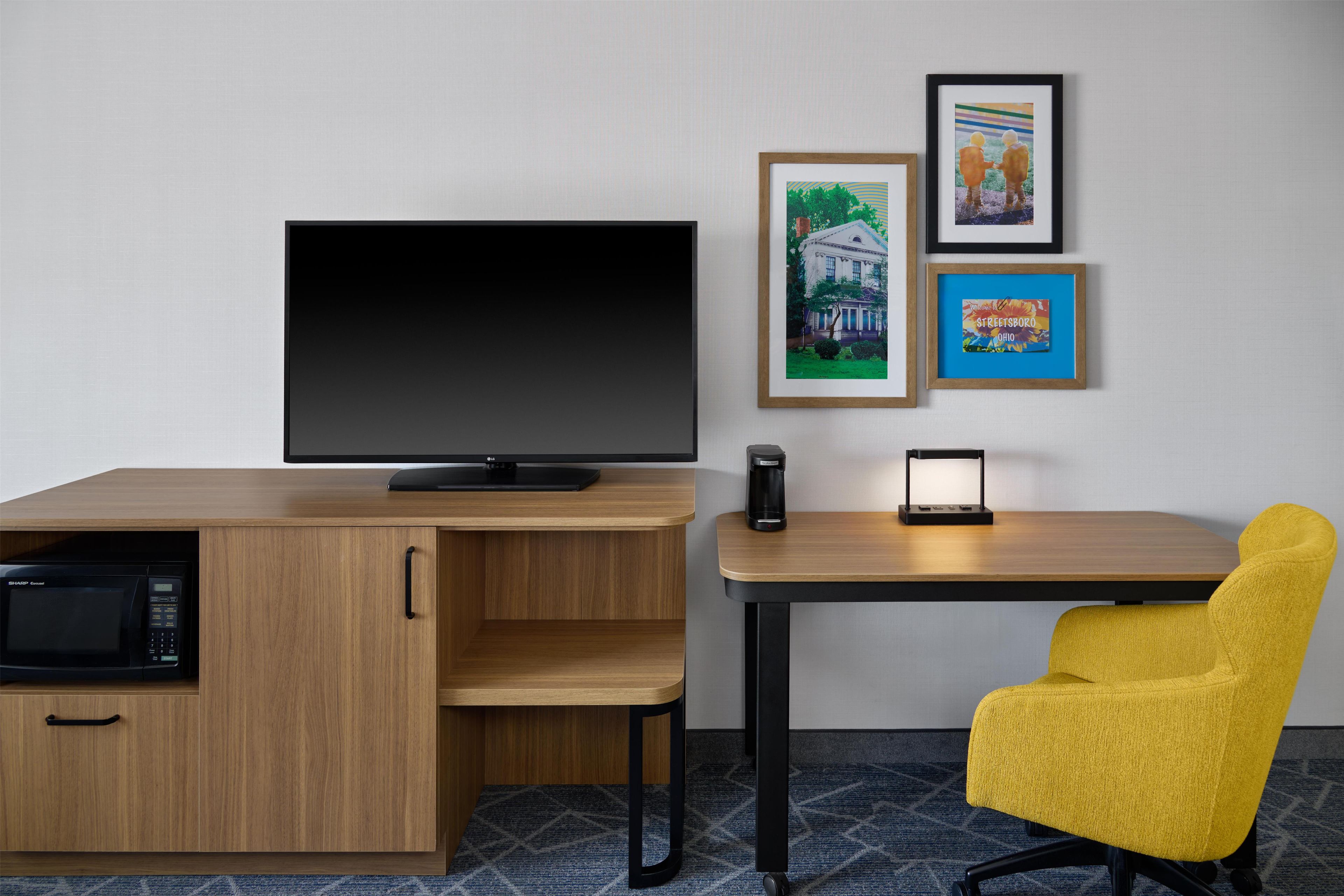A hotel room with a wooden desk, a flat-screen TV, a yellow chair, and framed pictures on the wall.