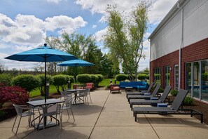 An outdoor patio with tables, chairs, and umbrellas, a brick building, and green landscaping. - Hampton Inn & Suites Cleveland Southeast/Streetsboro (Streetsboro)