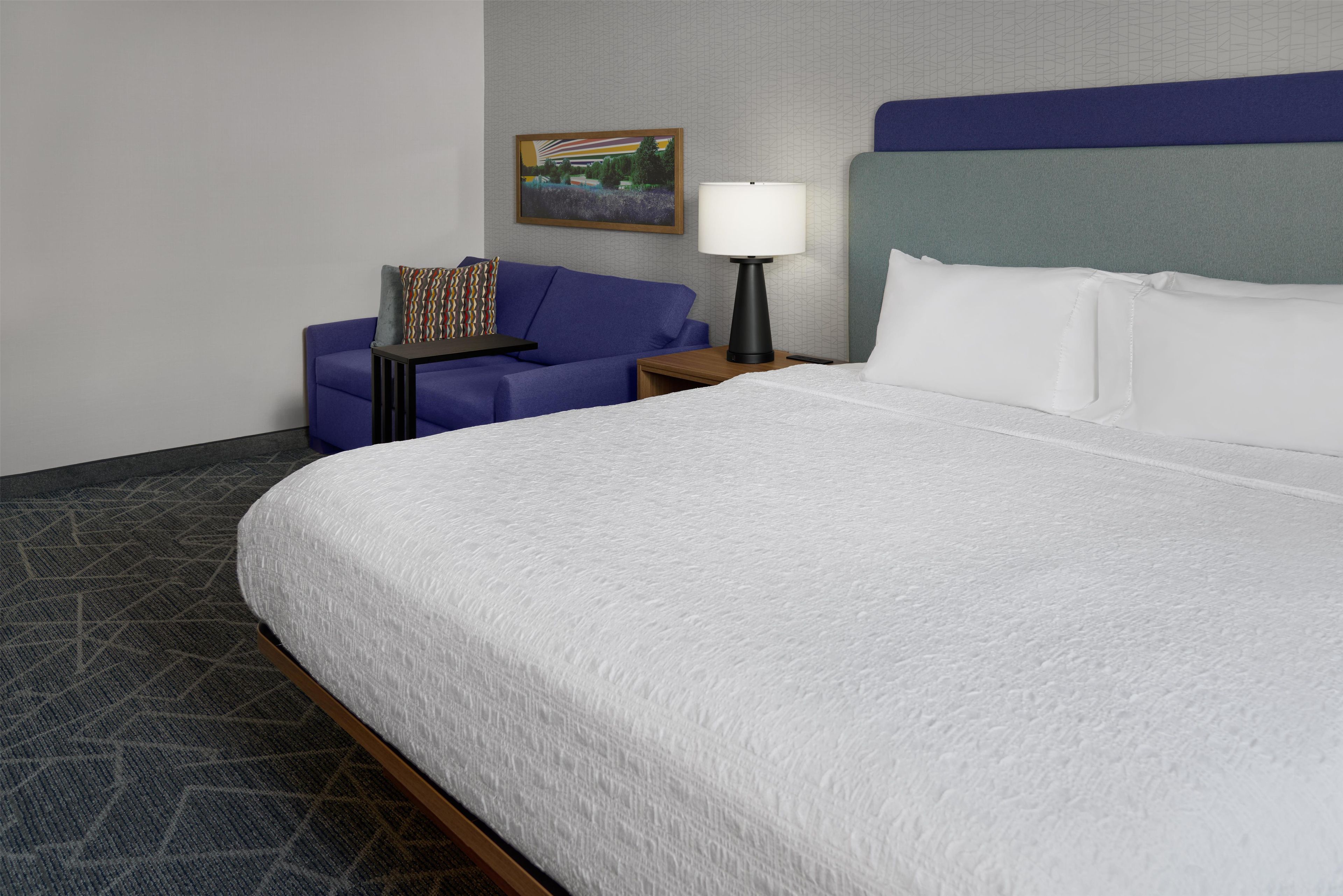 A hotel room with a large bed, a blue sofa, a small table, a lamp, and a painting on the wall.