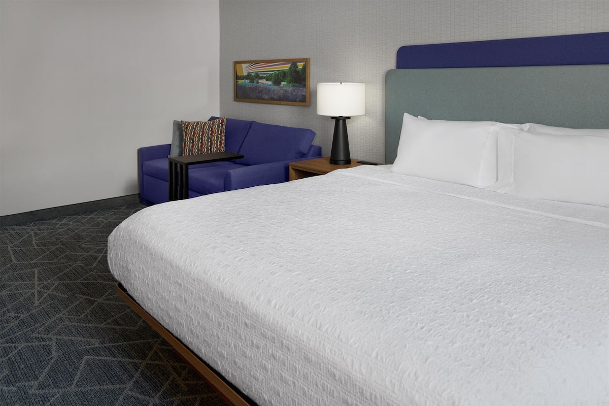 A hotel room with a large bed, a blue sofa, a small table, a lamp, and a painting on the wall.