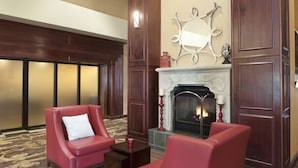 A hotel lobby with two red leather armchairs, a small table, a fireplace, and a decorative mirror above it.