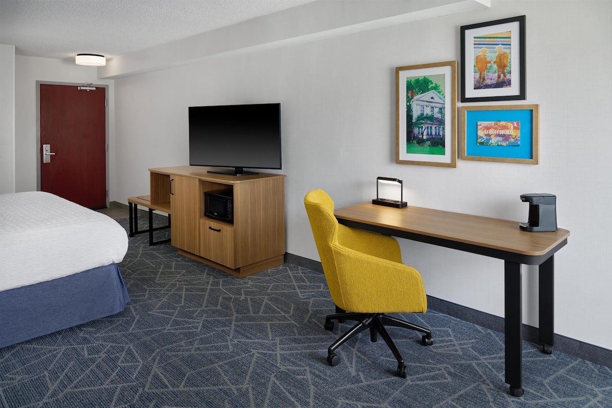 A hotel room with a bed, a desk with a chair, a television, and framed pictures on the wall.