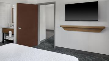 Habitación de hotel con una cama, un televisor montado en la pared, un estante de madera, un baño con espejo y una puerta que da a otra habitación.