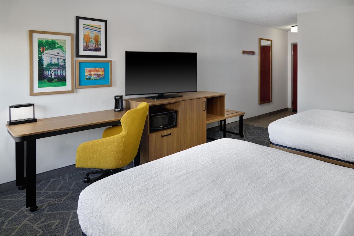 A hotel room with a desk, a television, two beds, a yellow chair, and framed pictures on the wall.