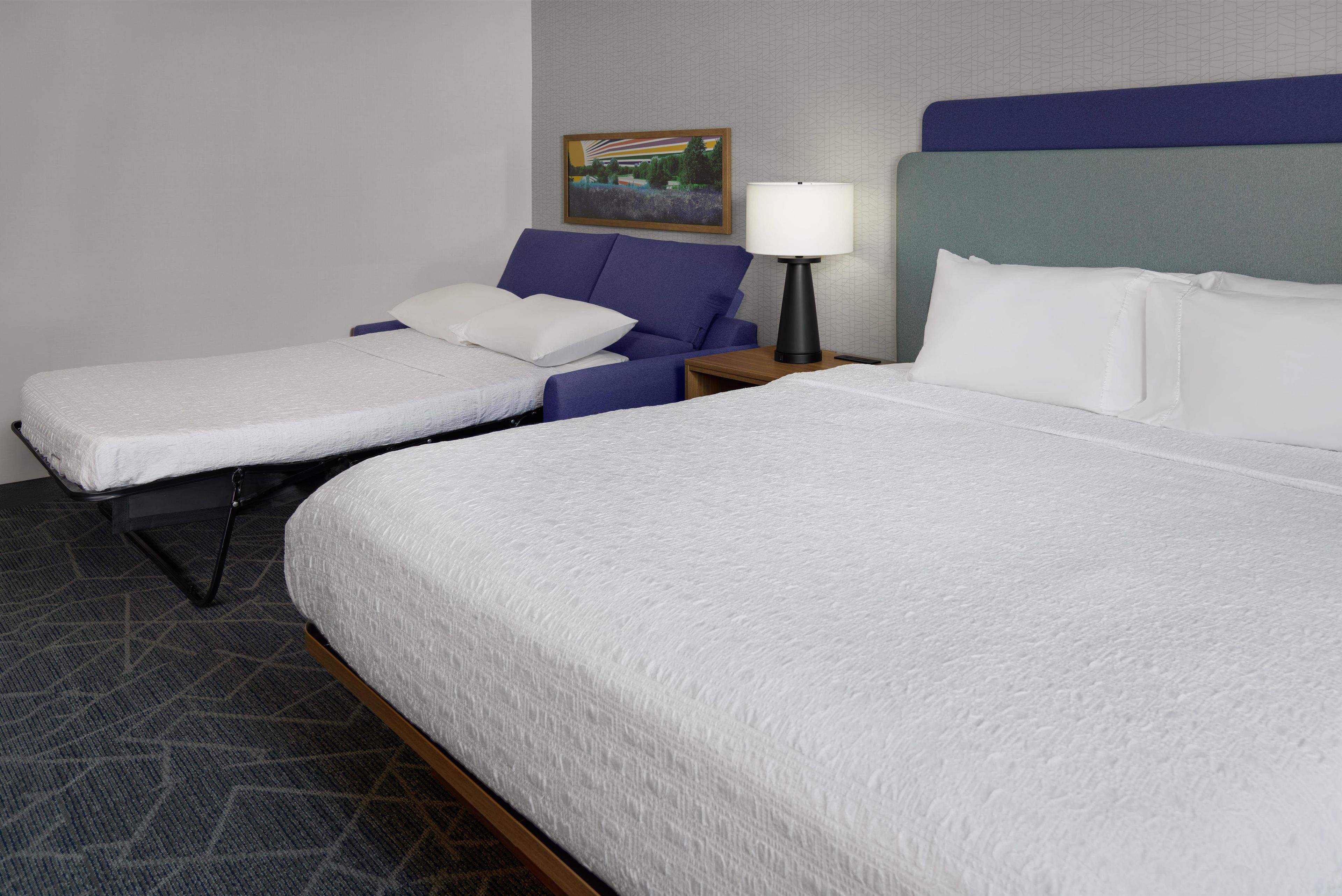 Una habitación de hotel con dos camas, una mesita de noche con lámpara y un cuadro en la pared.