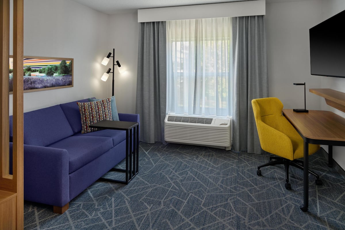 A hotel room with a blue sofa, a yellow chair, a small table, a mounted TV, and a large window with curtains.