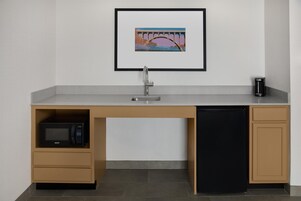 A compact kitchen with a microwave, sink, and dishwasher, a framed picture above the sink, and a small trash bin to the side.