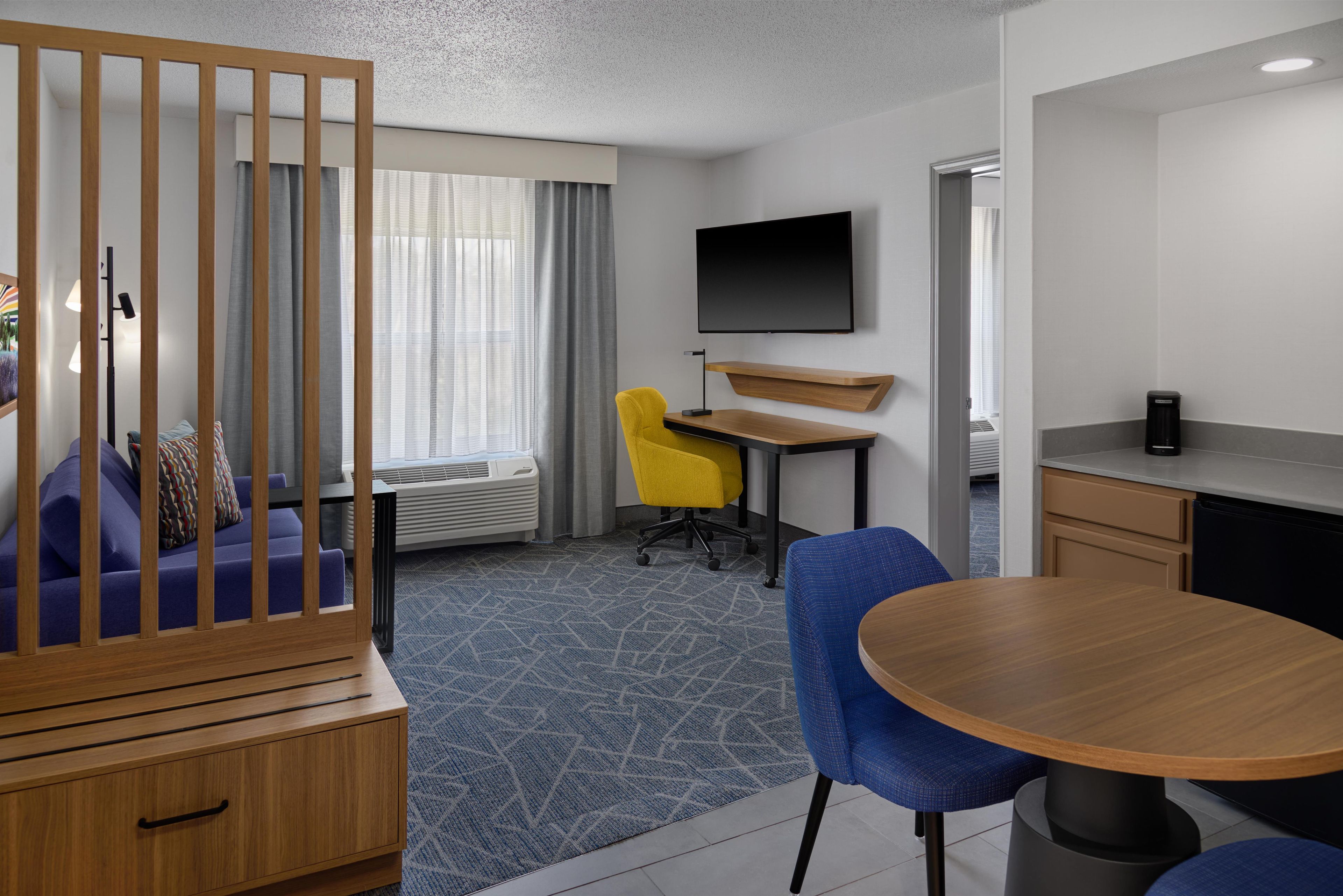 A hotel room with a wooden partition, a sofa, a desk with a chair, a TV mounted on the wall, and a dining table.