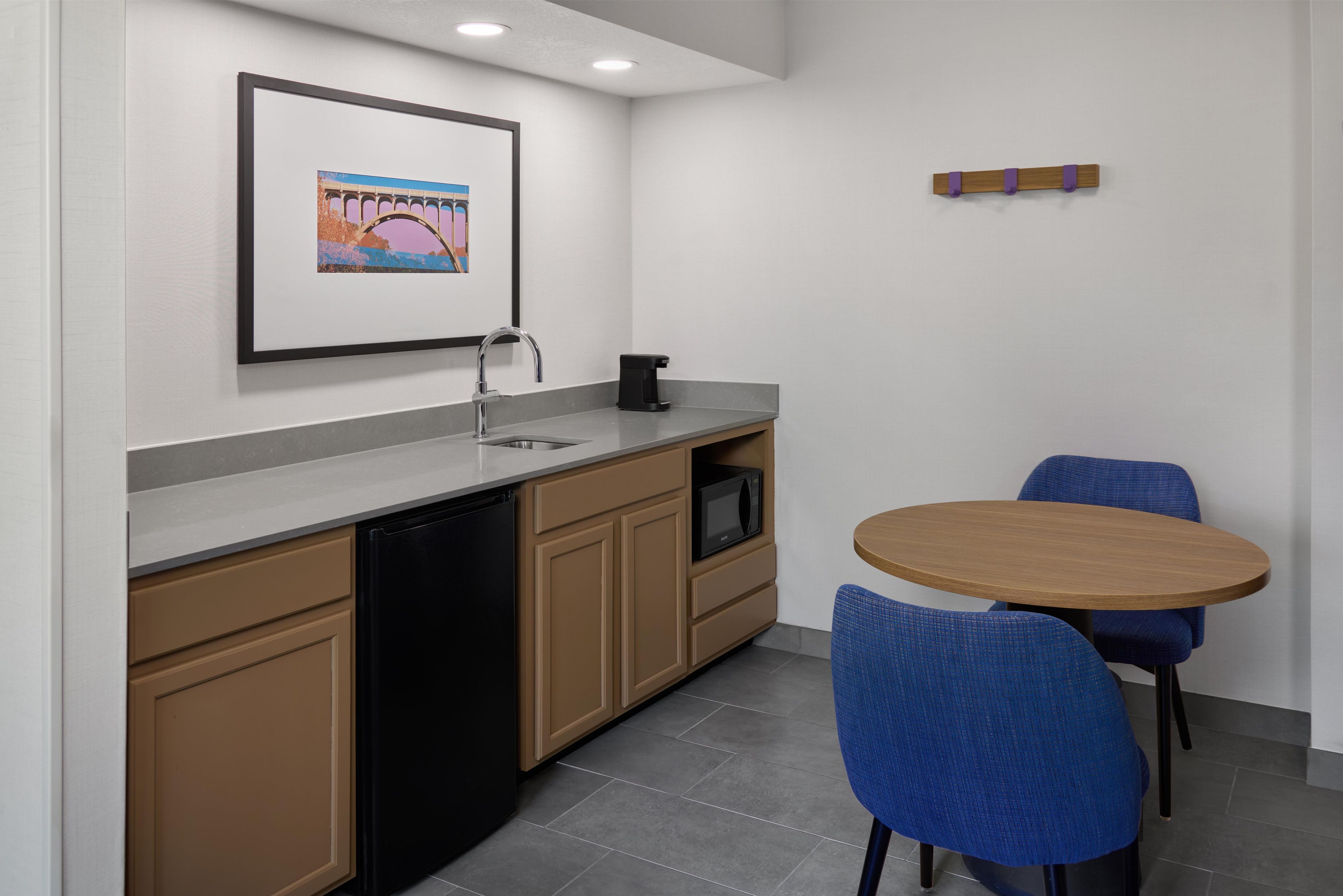 A modern kitchen with a dishwasher, microwave, and coffee maker. There is a framed picture on the wall and a small table with chairs.