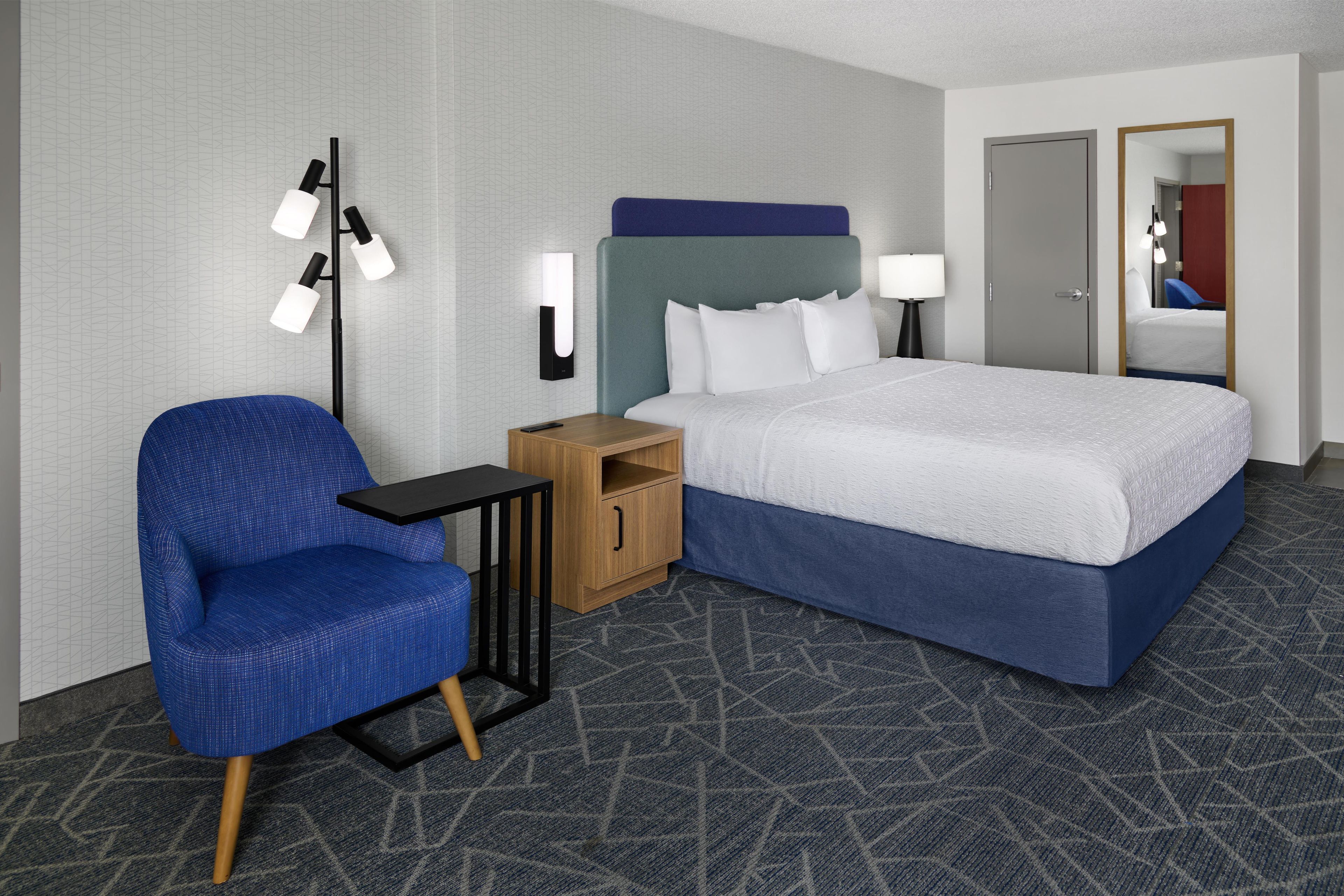 A hotel room with a bed, a chair, a nightstand, a lamp, and a mirror.