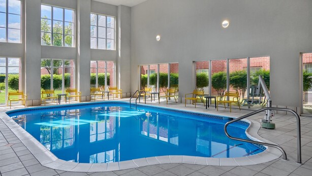 An indoor swimming pool with clear glass walls, yellow chairs, and a brick building in the background.