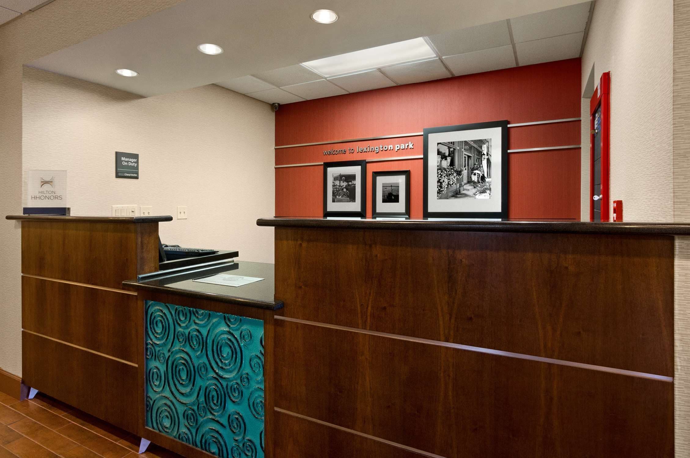 A reception desk with a wooden finish and a teal patterned panel, a red wall with a welcome sign, and framed black and white photographs.