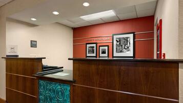 A reception desk with a wooden finish and a teal patterned panel, a red wall with a welcome sign, and framed black and white photographs.