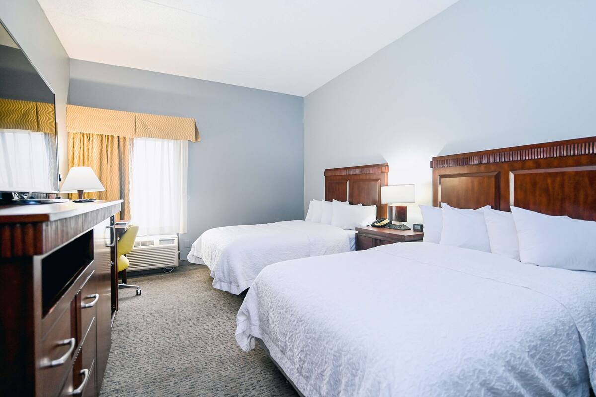A hotel room with two beds, a desk with a lamp, a chair, and a dresser.