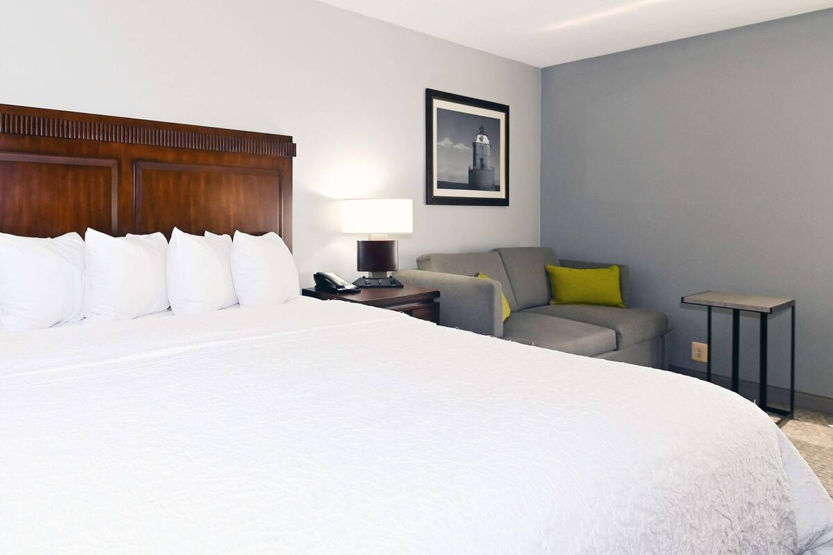 A hotel room with a large bed, a desk with a lamp, a grey sofa, and a small table.