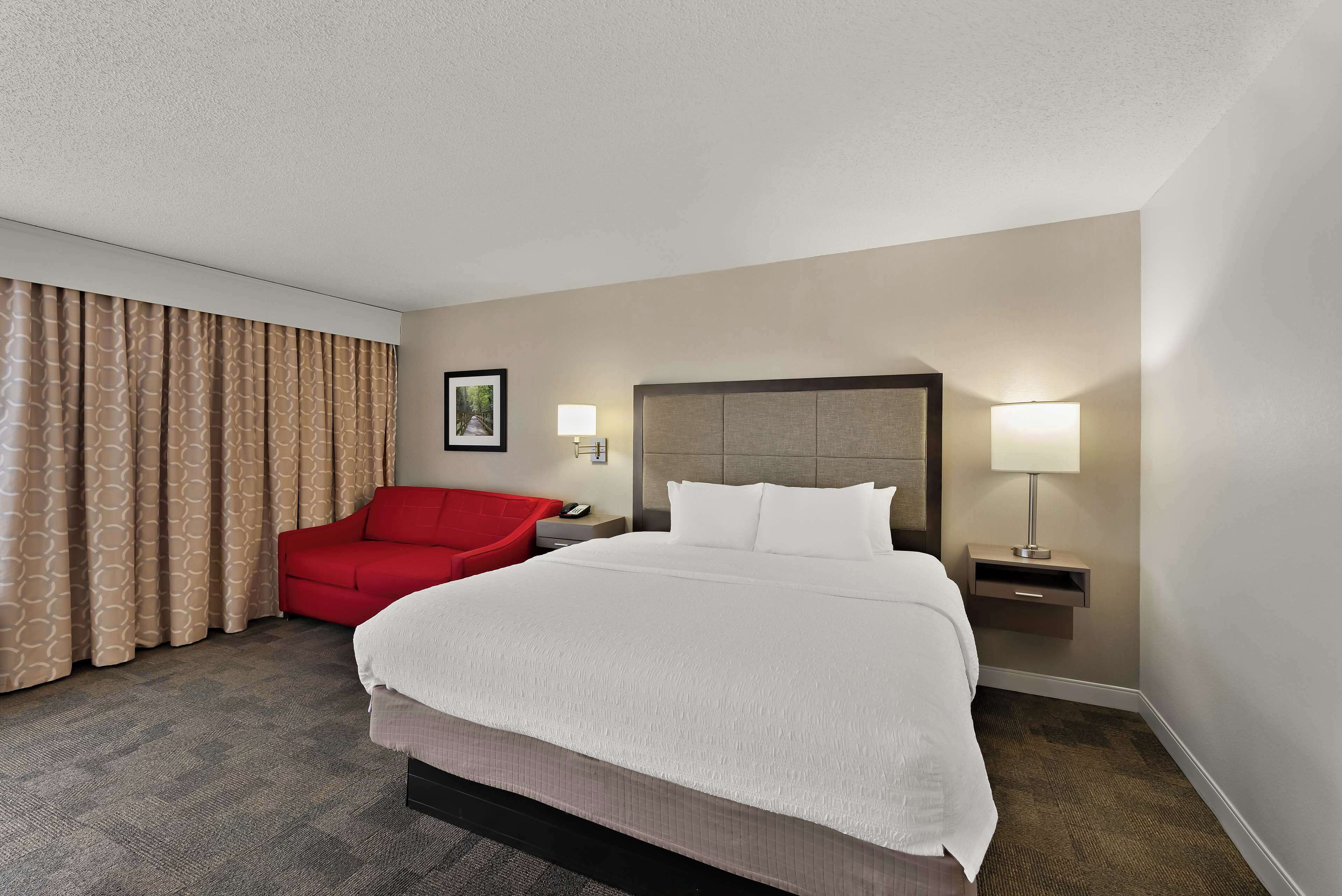Una habitación de hotel con una cama grande, una silla roja, una mesita de noche con una lámpara y un cuadro en la pared.