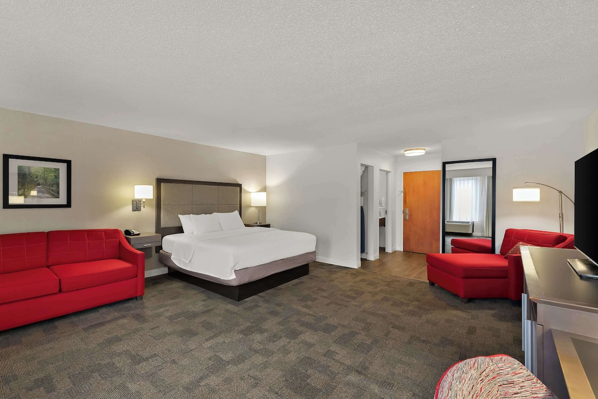 A hotel room with a bed, a red sofa, a red armchair, a desk, and a mirror.