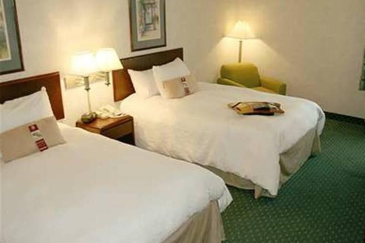 Ein Hotelzimmer mit zwei Betten, einem Nachttisch mit Lampe, einem Sessel und einem gerahmten Bild an der Wand.