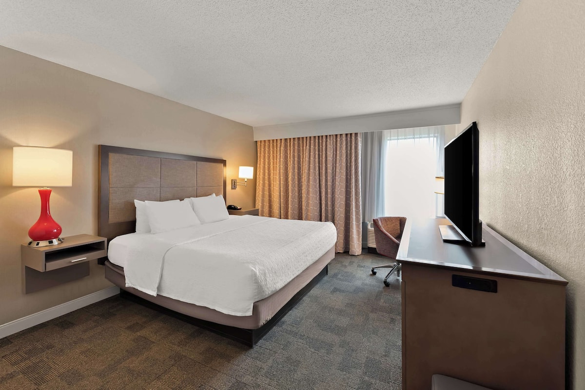 A hotel room with a large bed, a TV on a stand, a desk with a lamp, and a window with curtains.