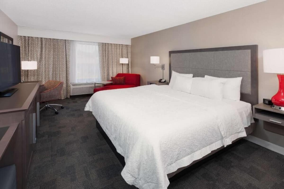 A hotel room with a large bed, a desk with a chair, a TV, and a red lamp.