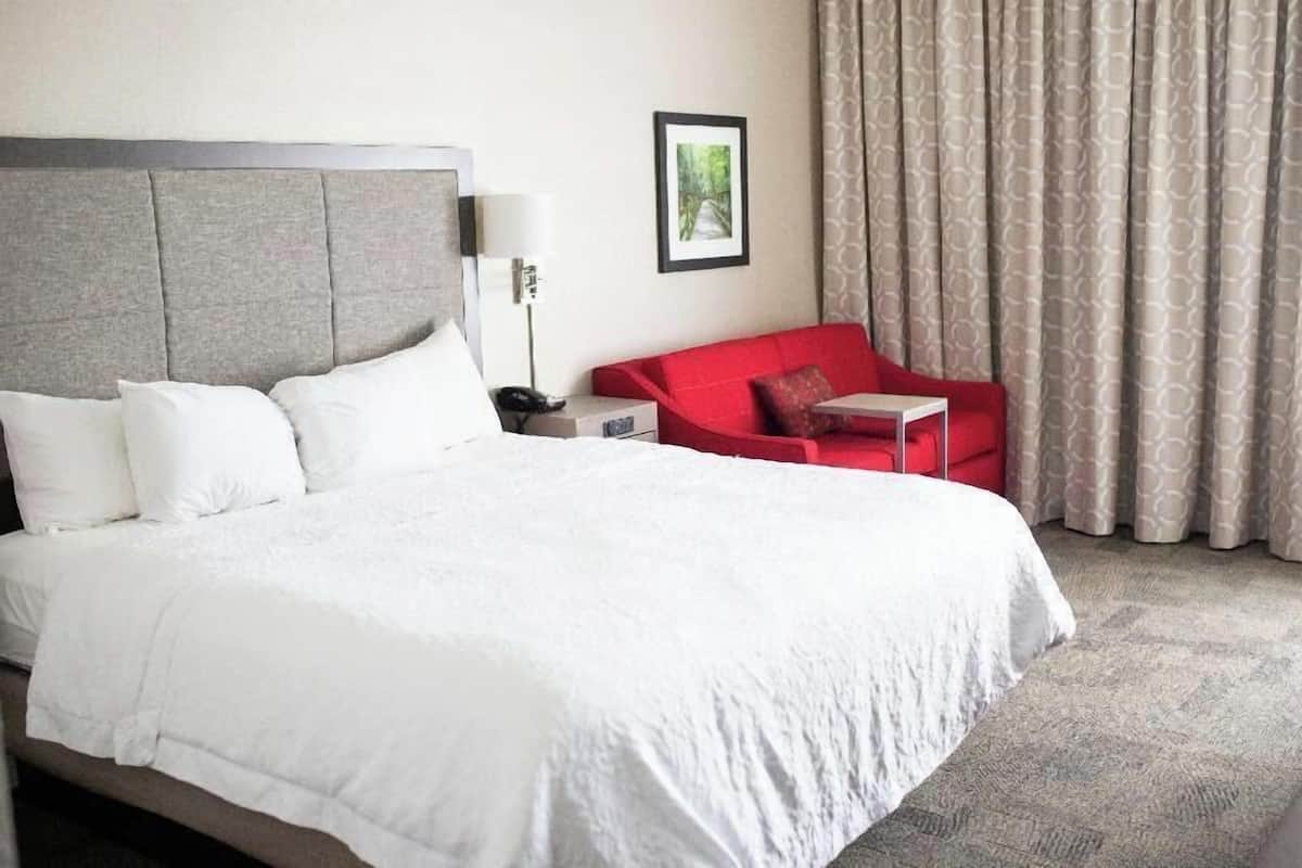 A hotel room with a large bed, a red armchair, a bedside table with a lamp, and a framed picture on the wall.
