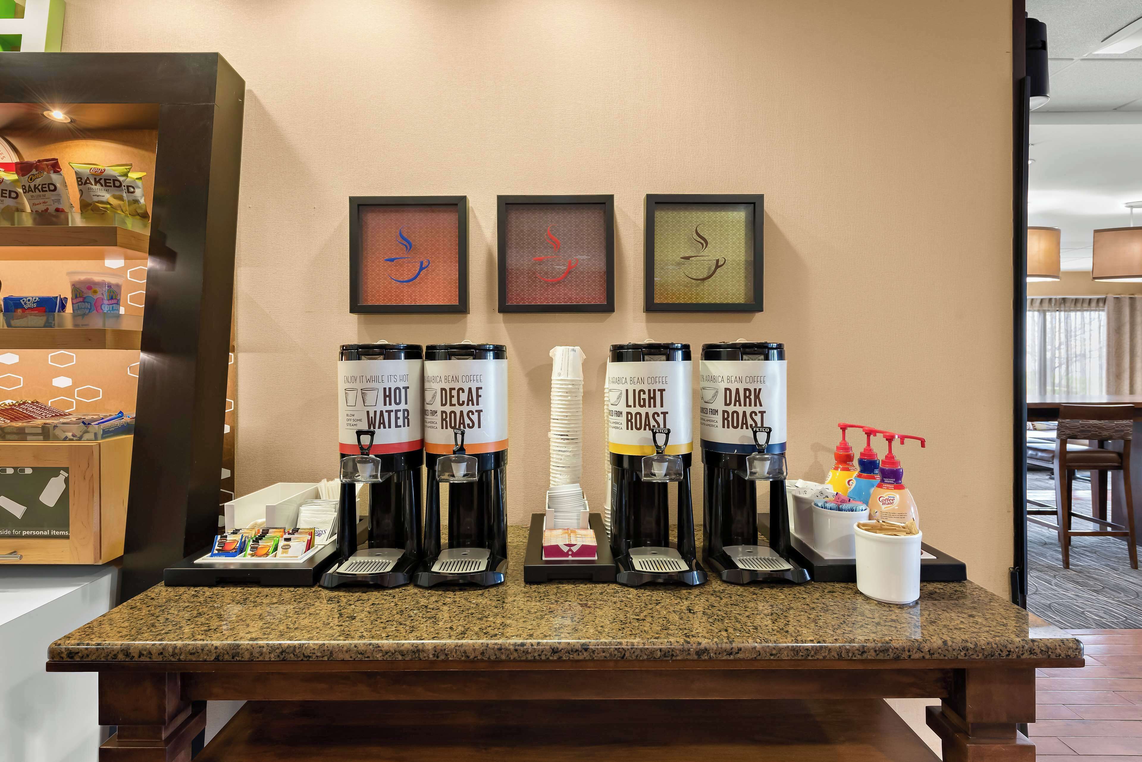 Una estación de café con cuatro dispensadores de café etiquetados como 'Hot Water', 'Decaf Roast', 'Light Roast' y 'Dark Roast'. También hay dispensadores de condimentos y una pequeña exhibición de bocadillos.