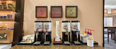 Una estación de café con cuatro dispensadores de café etiquetados como 'Hot Water', 'Decaf Roast', 'Light Roast' y 'Dark Roast'. También hay dispensadores de condimentos y una pequeña exhibición de bocadillos.