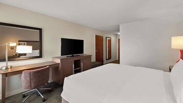Quarto de hotel com cama, escrivaninha com televisão, cadeira e espelho.