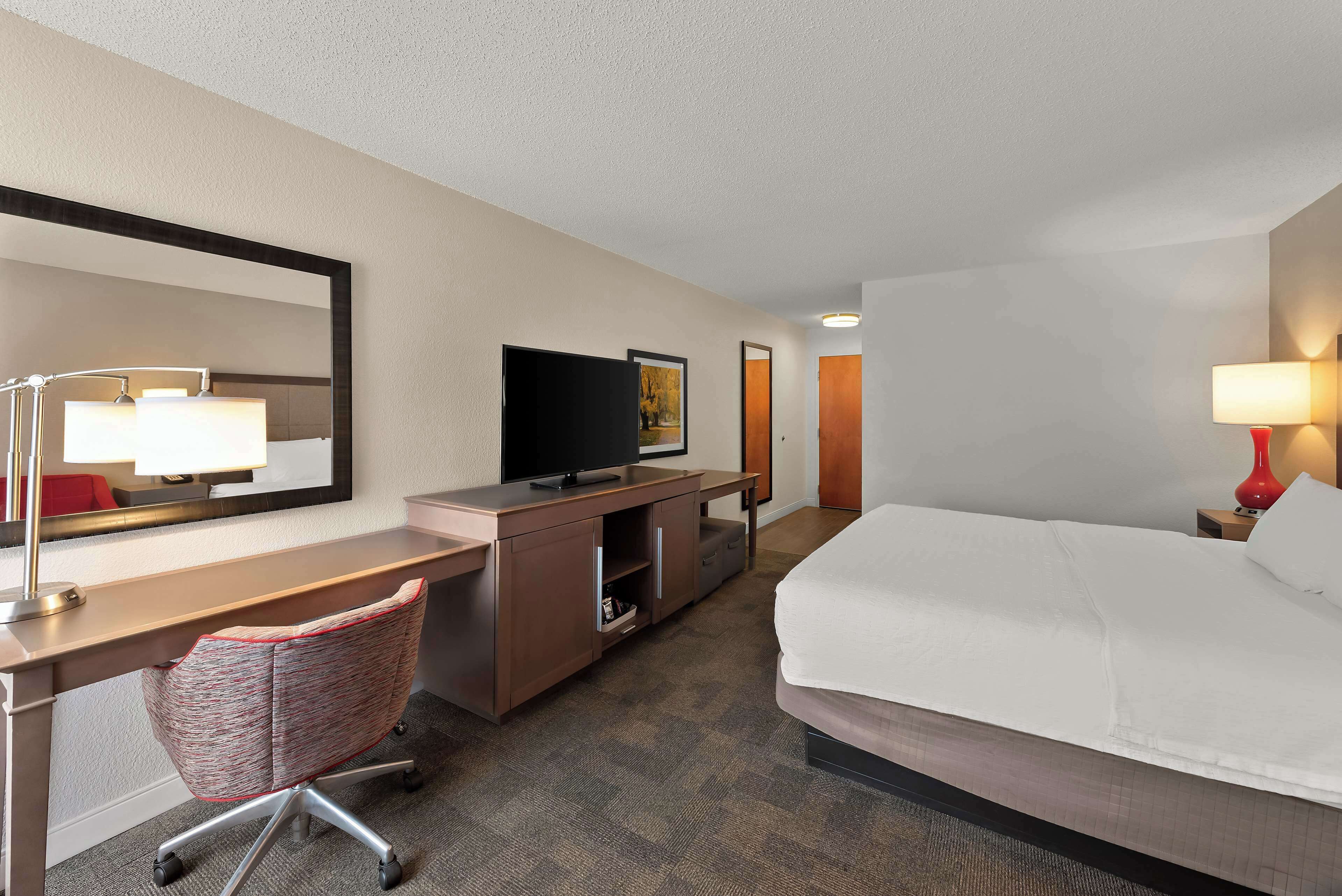Una habitación de hotel con cama, escritorio, silla, televisor y un espejo grande.