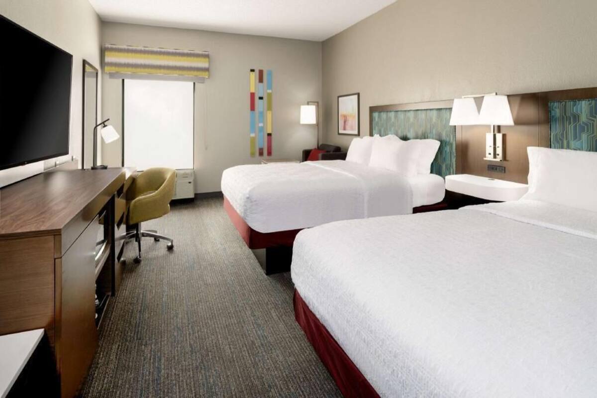 A hotel room with two beds, a desk, a chair, a TV, and a window with blinds.